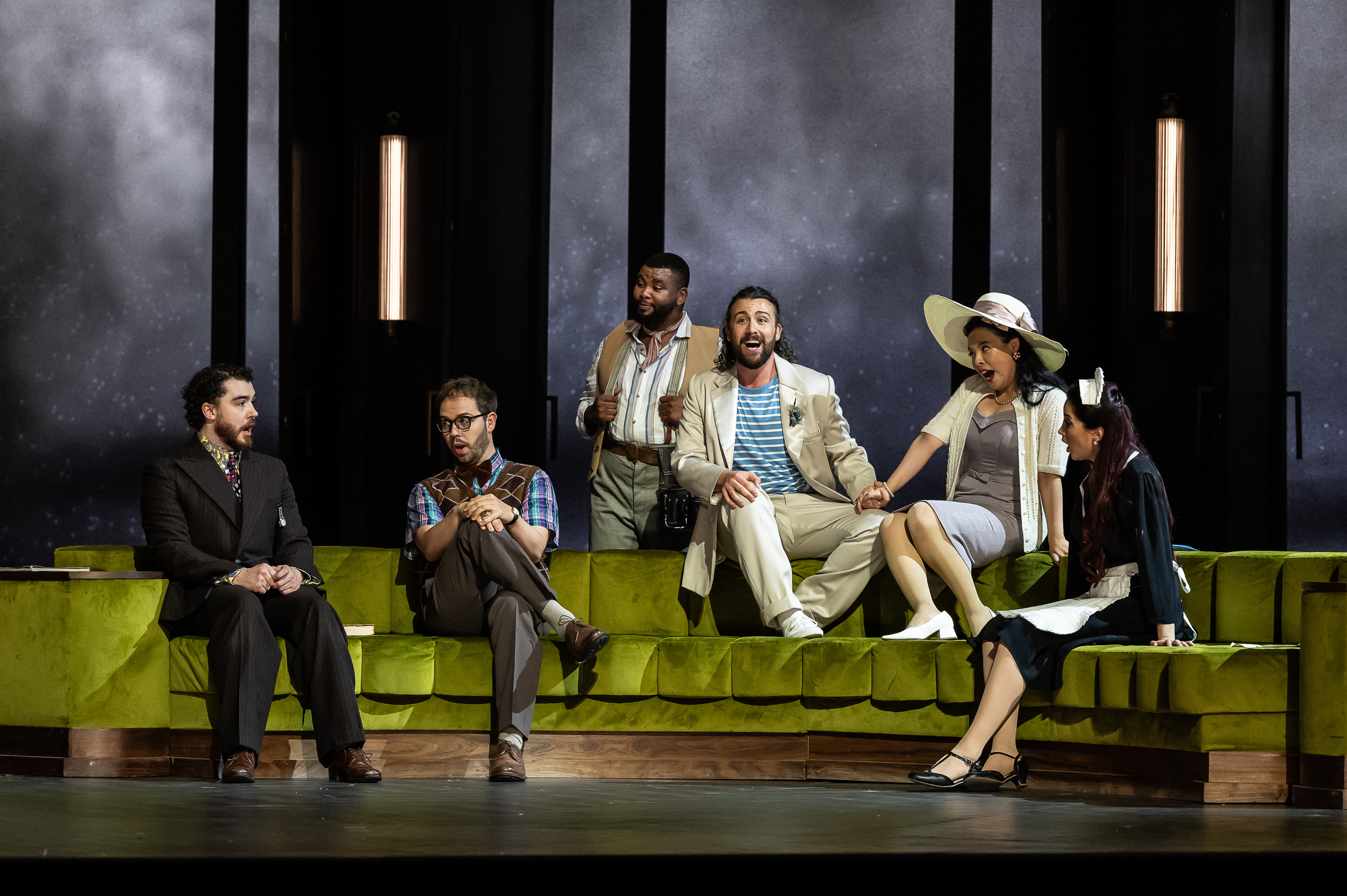 A group of Jette Parker Singers sit together on a green velvet couch against a plain grey backdrop. The men wear suits in different colours, while one woman wears a large white hat and another is dressed in a maid’s outfit.