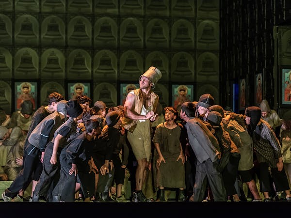 A performer wearing a bucket on their head and ragged clothes stands in the centre surrounded by dozens of small children, also wearing ragged and dirty clothes.