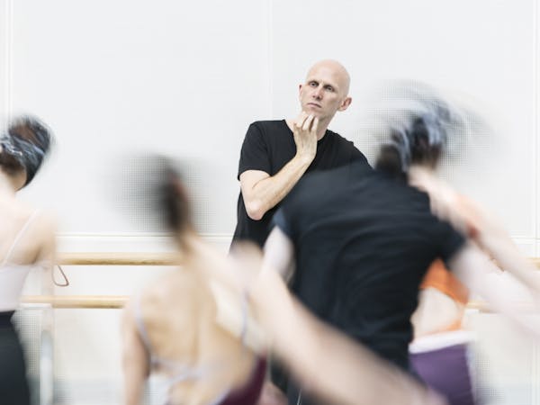 Resident Choreographer of The Royal Ballet Wayne McGregor stands with his right hand to his chin in the centre of a dance rehearsal studio. Dancers around him are artistically blurred as if in movement.