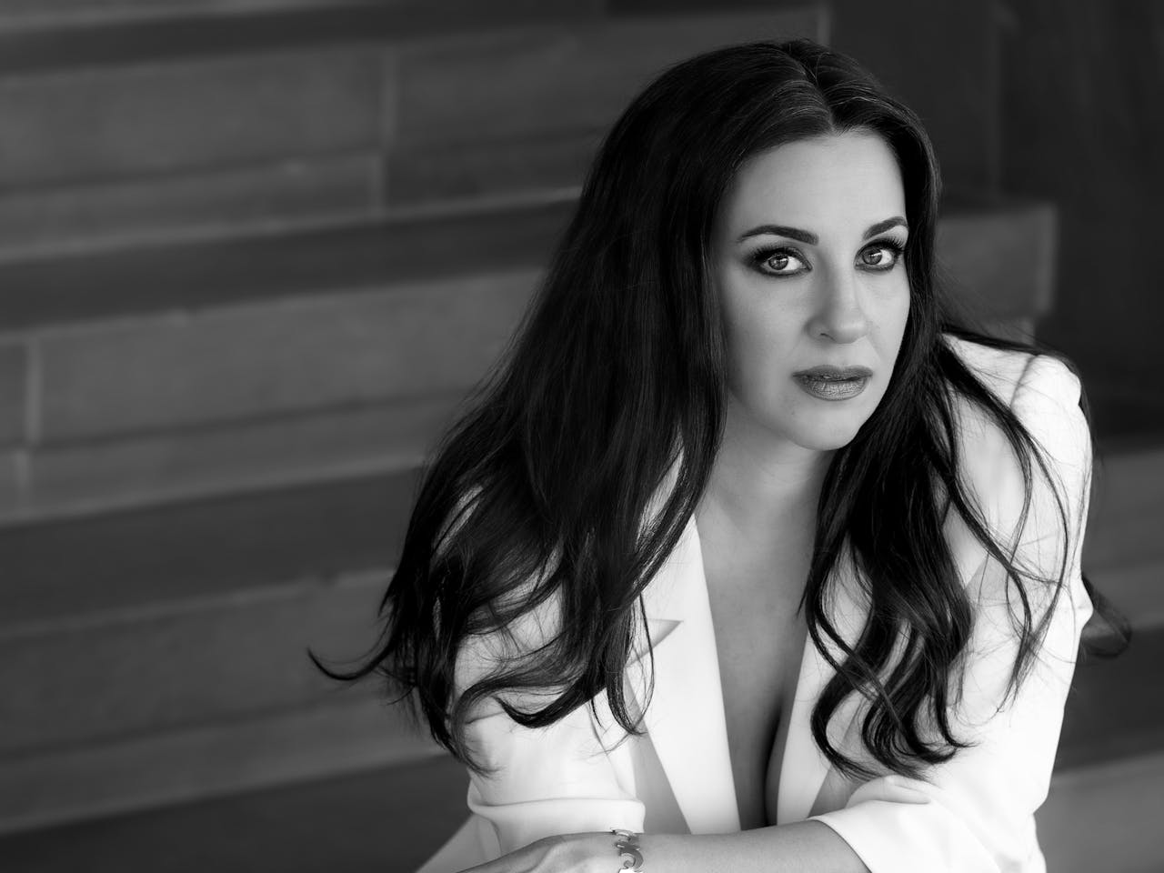 A black and white headshot of the opera singer Joyce El-Khoury. She sits against a brick wall wearing a white suit jacket.