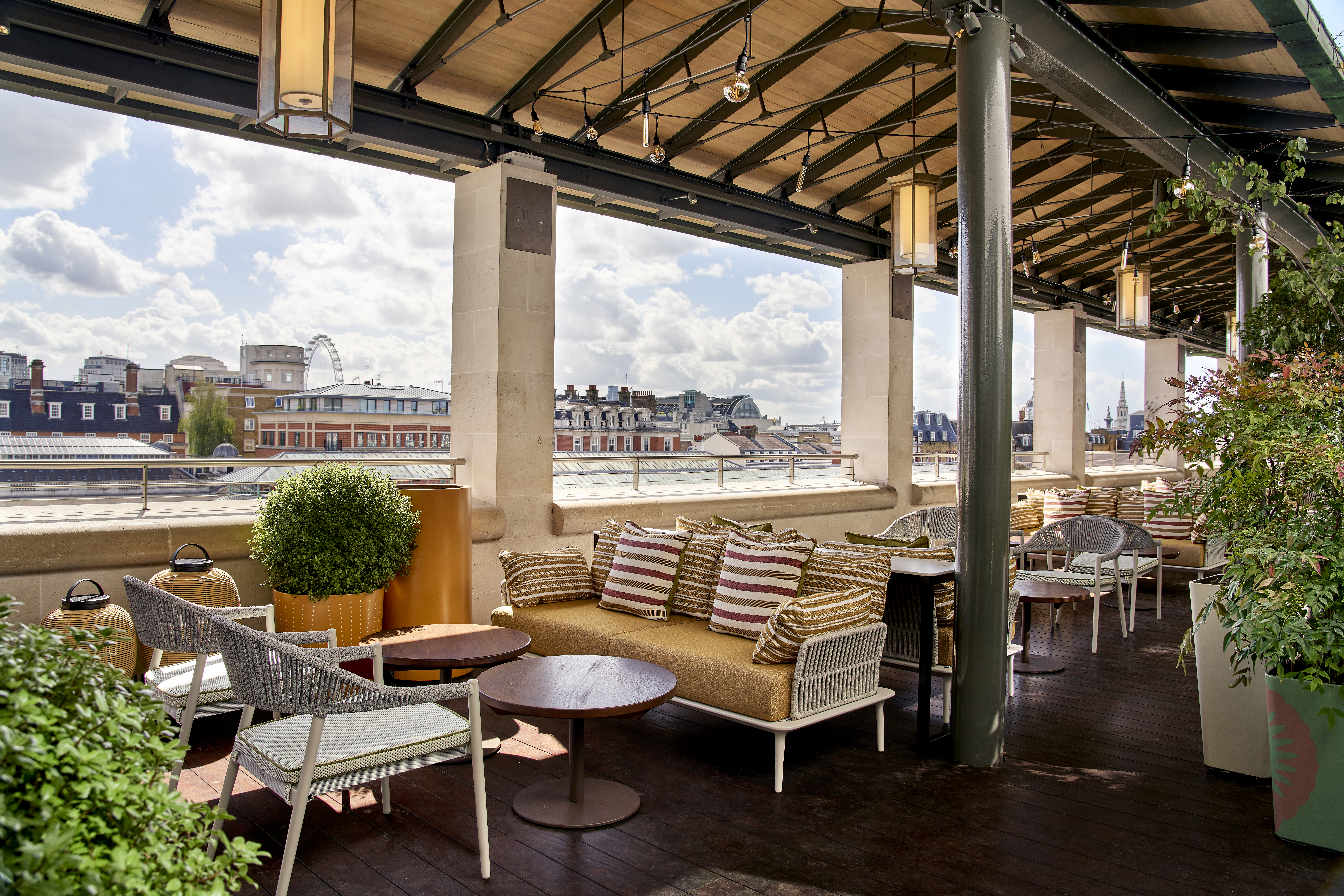 A day-time view of Bar Cicoria's covered rooftop terrace, featuring plush green chairs and yellow sofas, wooden tables, soft lighting and greenery. In the background is the London city skyline, including landmarks like the London Eye. 