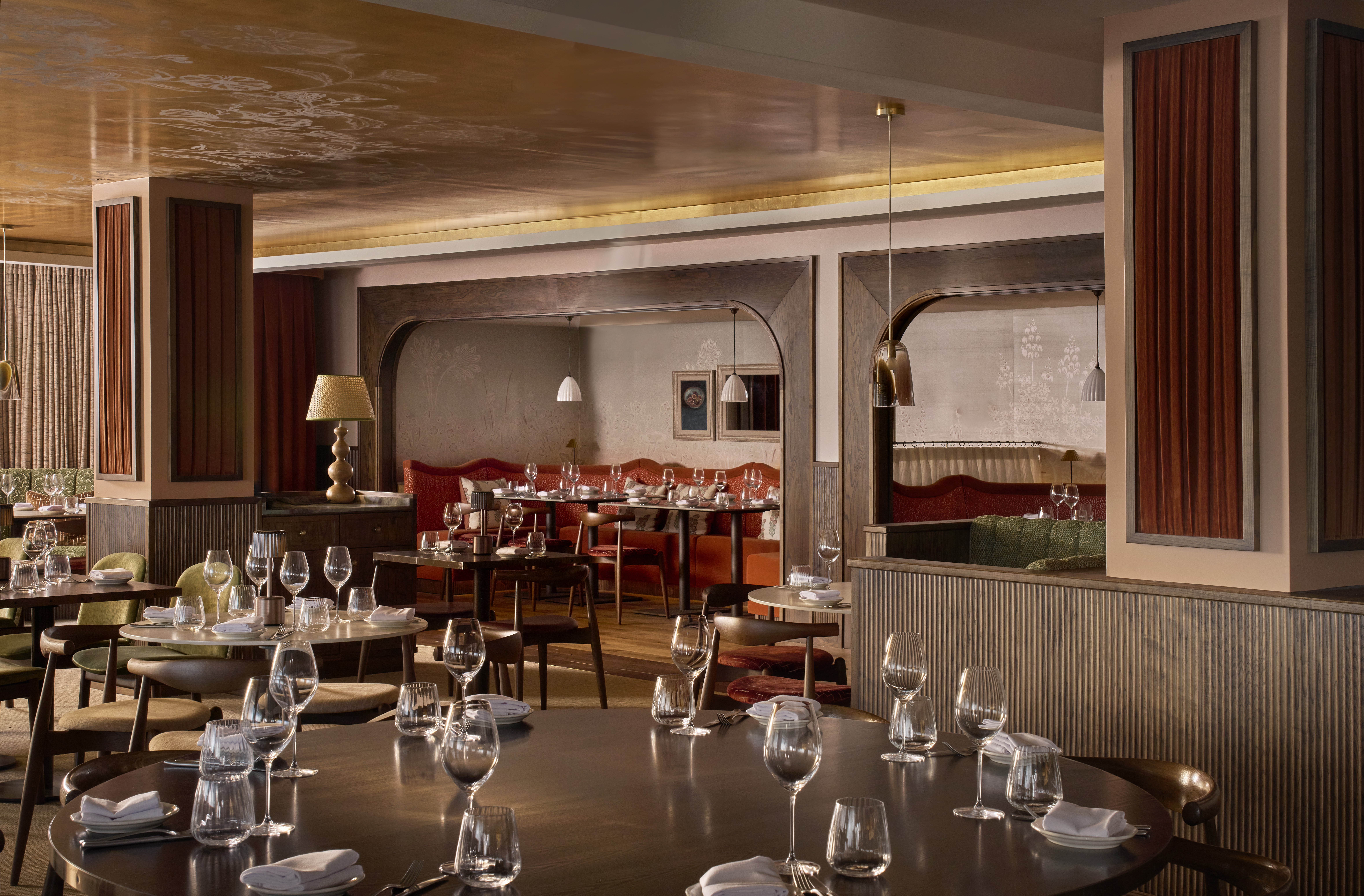 An interior view of Cicoria, Angela Hartnett's restaurant at the Royal Opera House. The space is decorated in a warm colour palette, including pink, deep orange and green tones alongside dark wooden panelling. Several wooden tables fill the room, each laid with plates and wine glasses. 