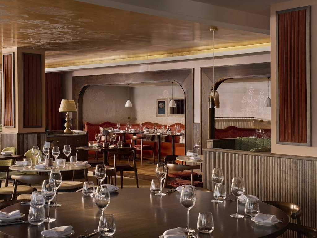 An interior view of Cicoria, Angela Hartnett's restaurant at the Royal Opera House. The space is decorated in a warm colour palette, including pink, deep orange and green tones alongside dark wooden panelling. Several wooden tables fill the room, each laid with plates and wine glasses.
