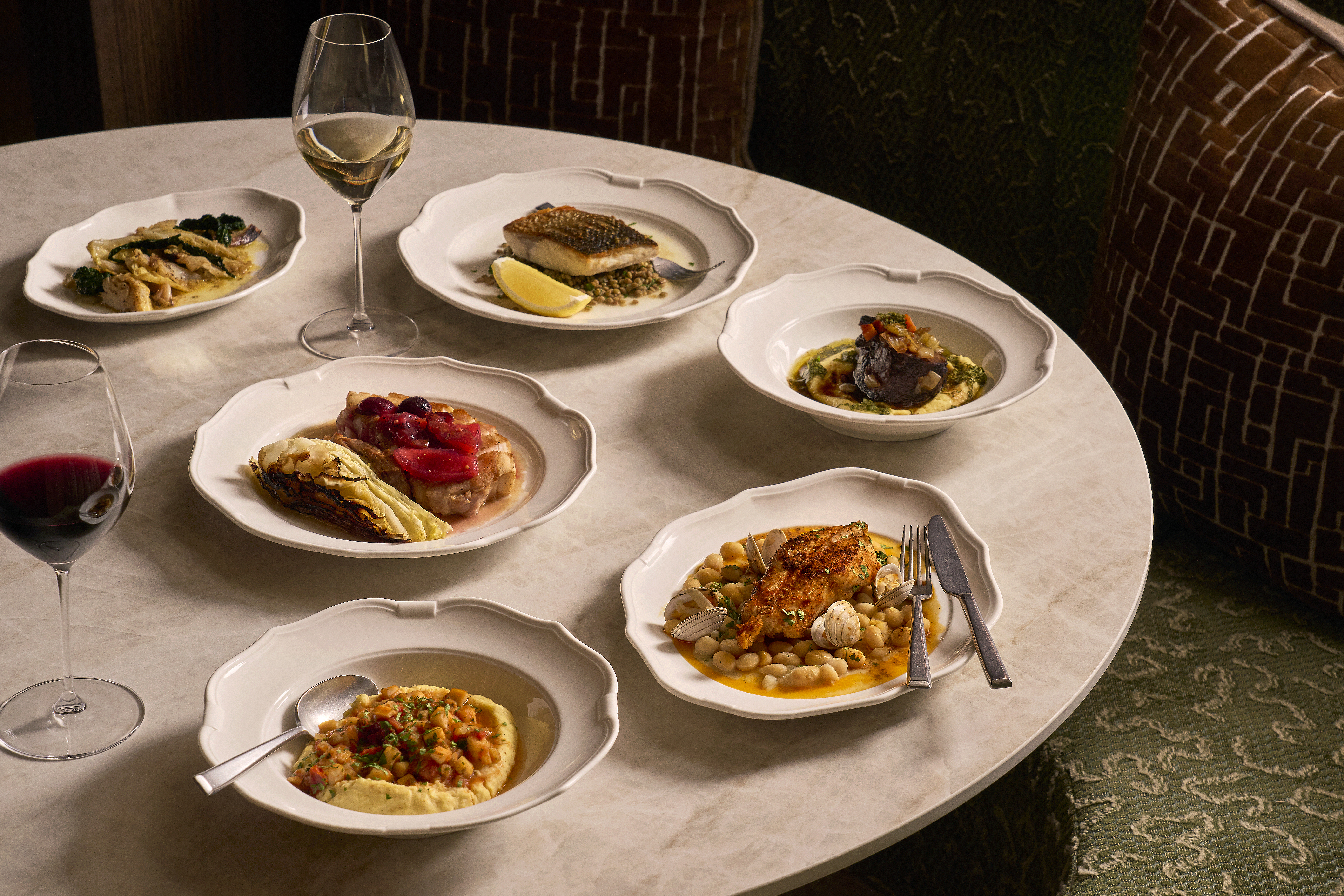 Plates of artfully styled food and glasses of red and white wine are arranged on a round marble table. The display contains menu items from Cicoria, Angela Hartnett's restaurant at the Royal Opera House.