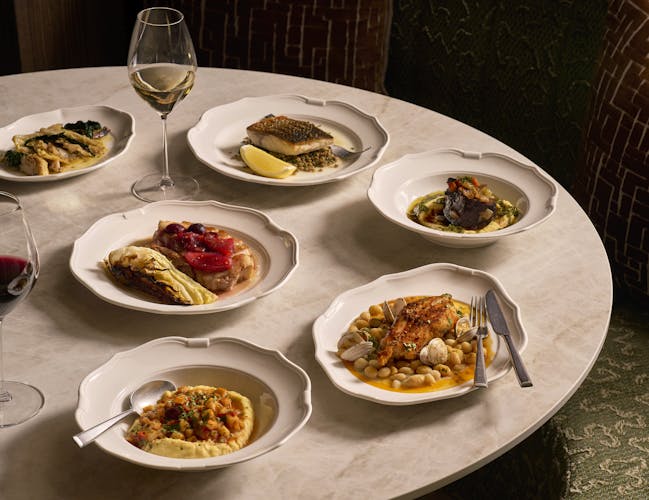 Plates of artfully styled food and glasses of red and white wine are arranged on a round marble table. The display contains menu items from Cicoria, Angela Hartnett's restaurant at the Royal Opera House.