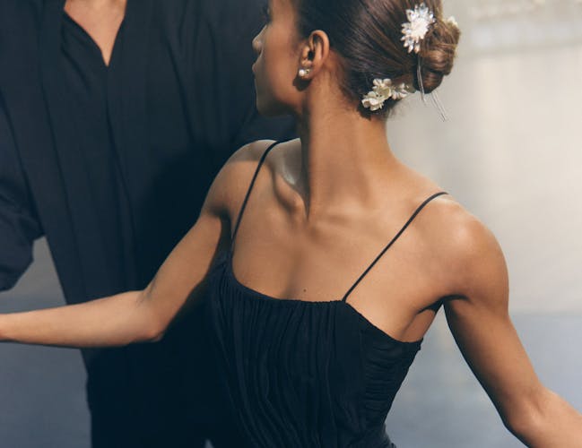 A woman in a black dress with white flowers in her hair holds hands with a man in a black suit.