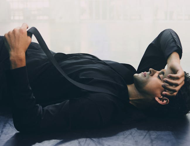 A man in a black shirt and tie lies on the floor with a hand covering his face.