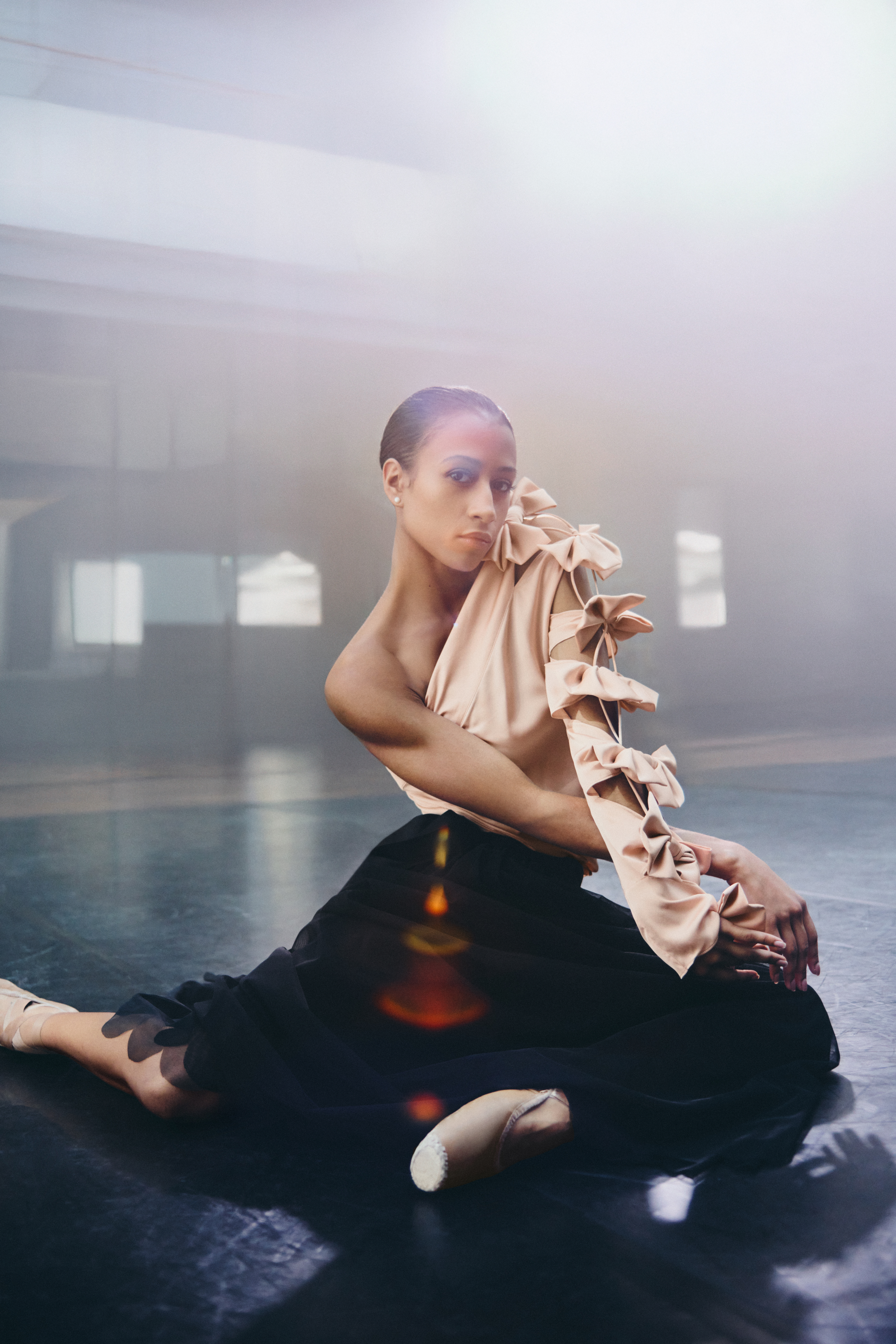 A woman in a pink satin shirt with bows and a black tulle skirt sits on the floor in a sunlit room.