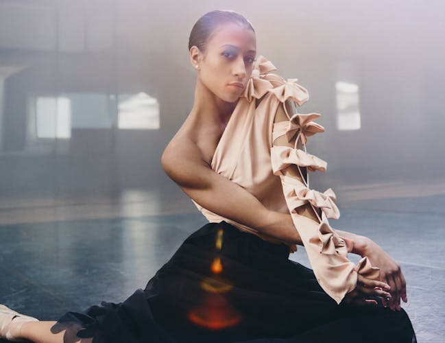 A woman in a pink satin shirt with bows and a black tulle skirt sits on the floor in a sunlit room.