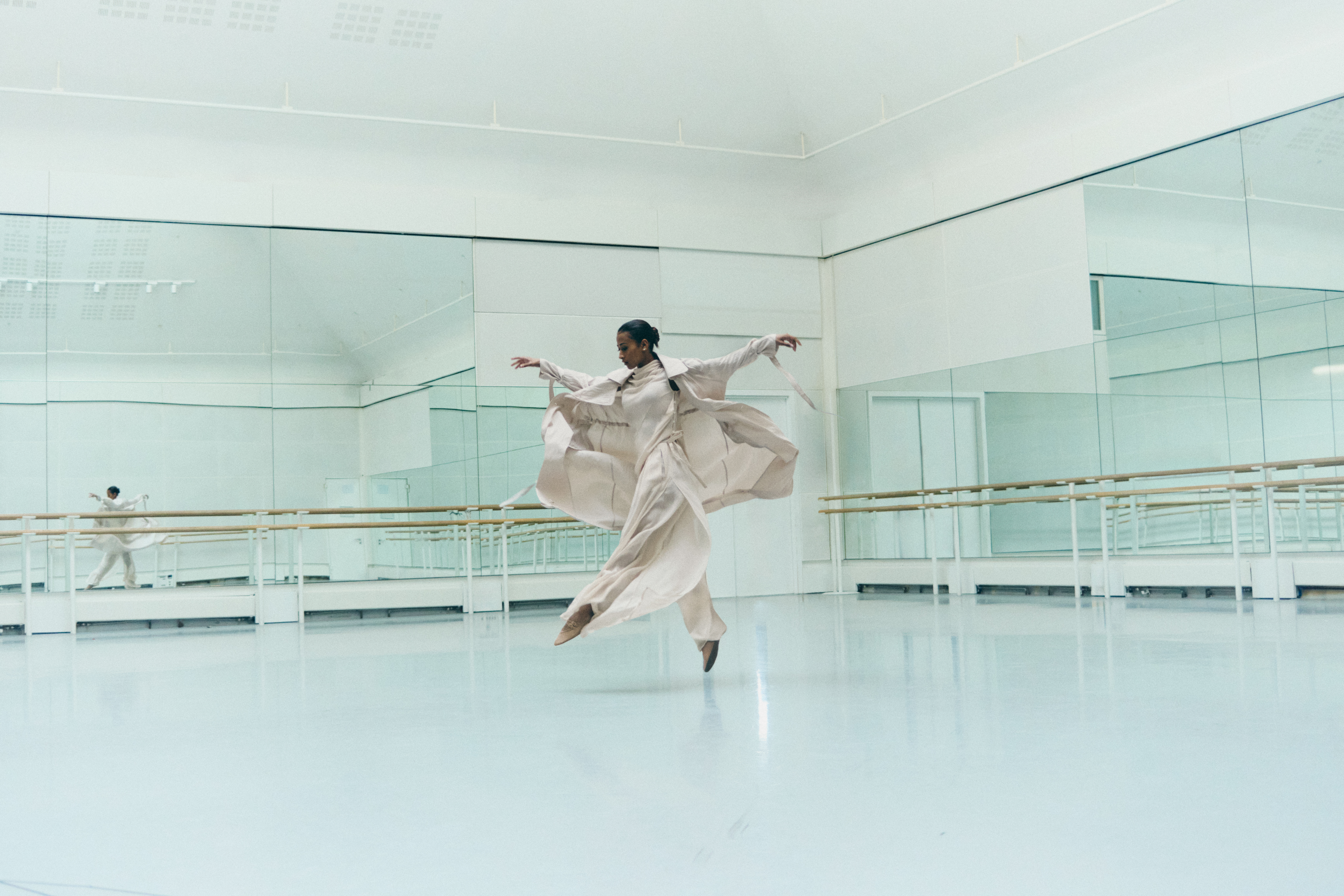 A women wearing loose cream clothing jumps in the centre of a dance studio.