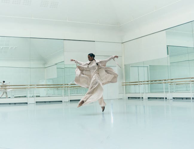 A women wearing loose cream clothing jumps in the centre of a dance studio.