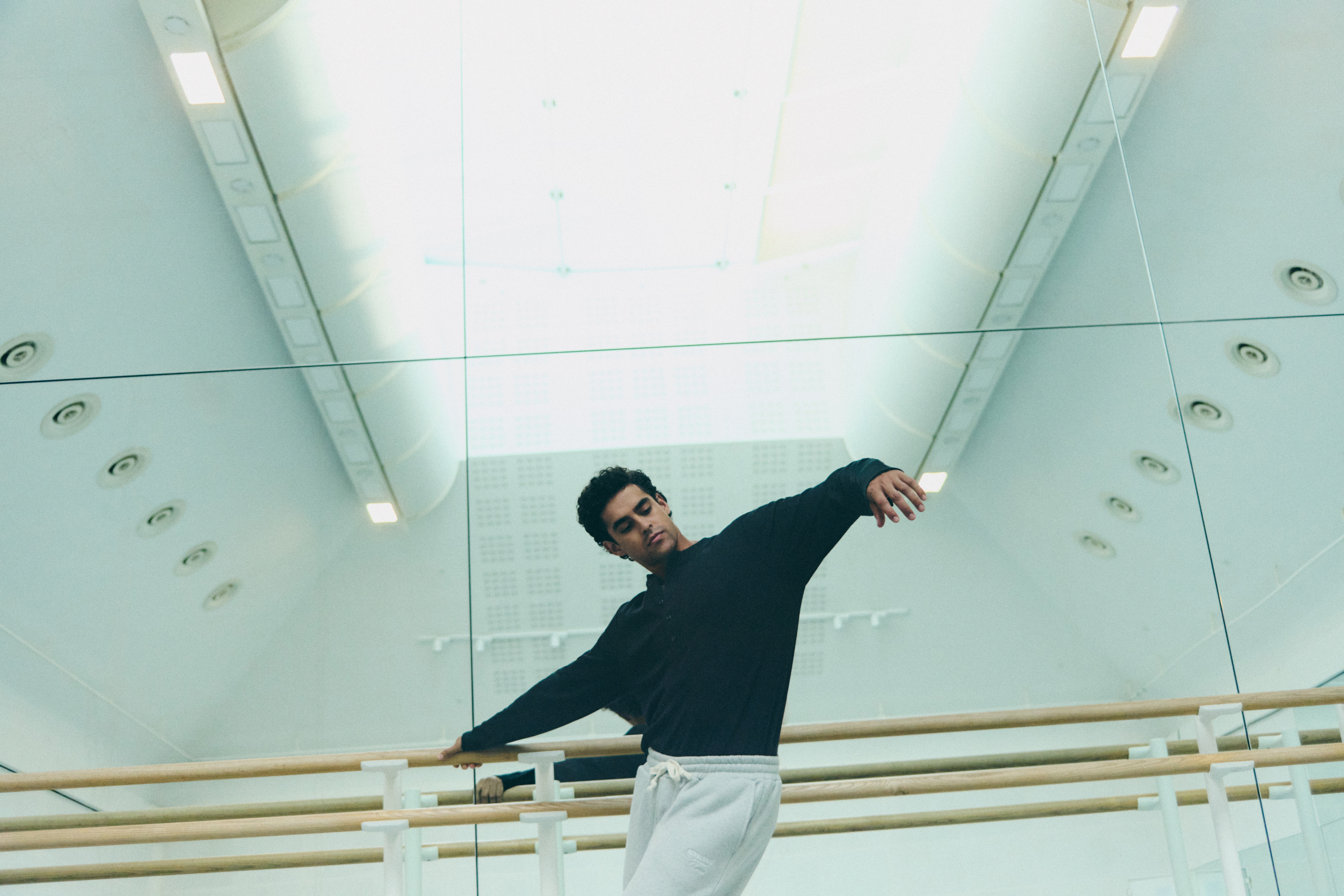 A man wearing a black jumper and grey joggers leans near a ballet barre in a dance studio