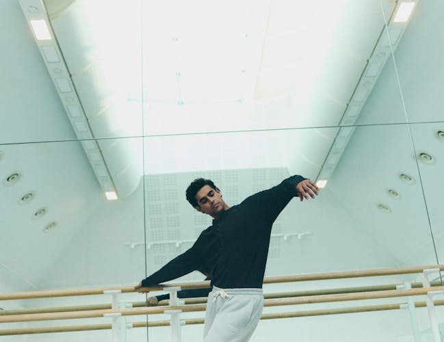 A man wearing a black jumper and grey joggers leans near a ballet barre in a dance studio