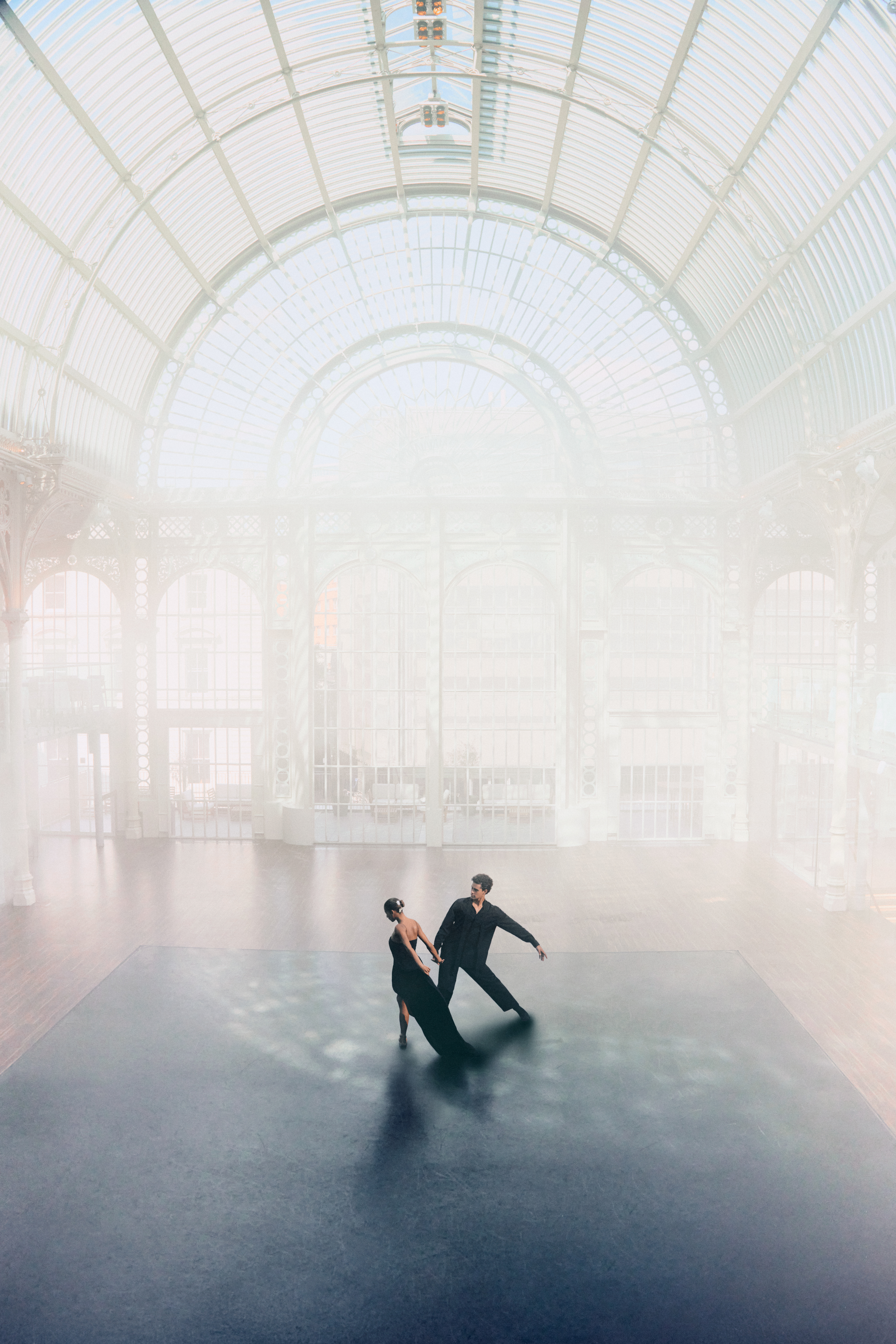 Two dancers wearing black pose together in a sunlit room.