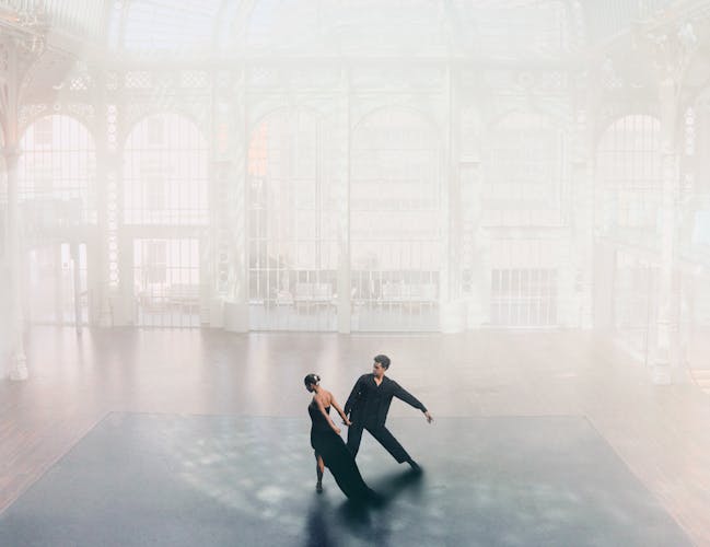 Two dancers wearing black pose together in a sunlit room.