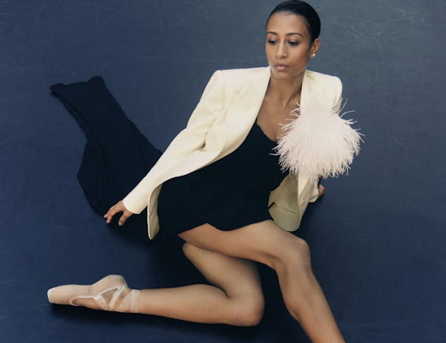A woman in a white blazer with a fringed embellishment and a black dress sits on the floor.
