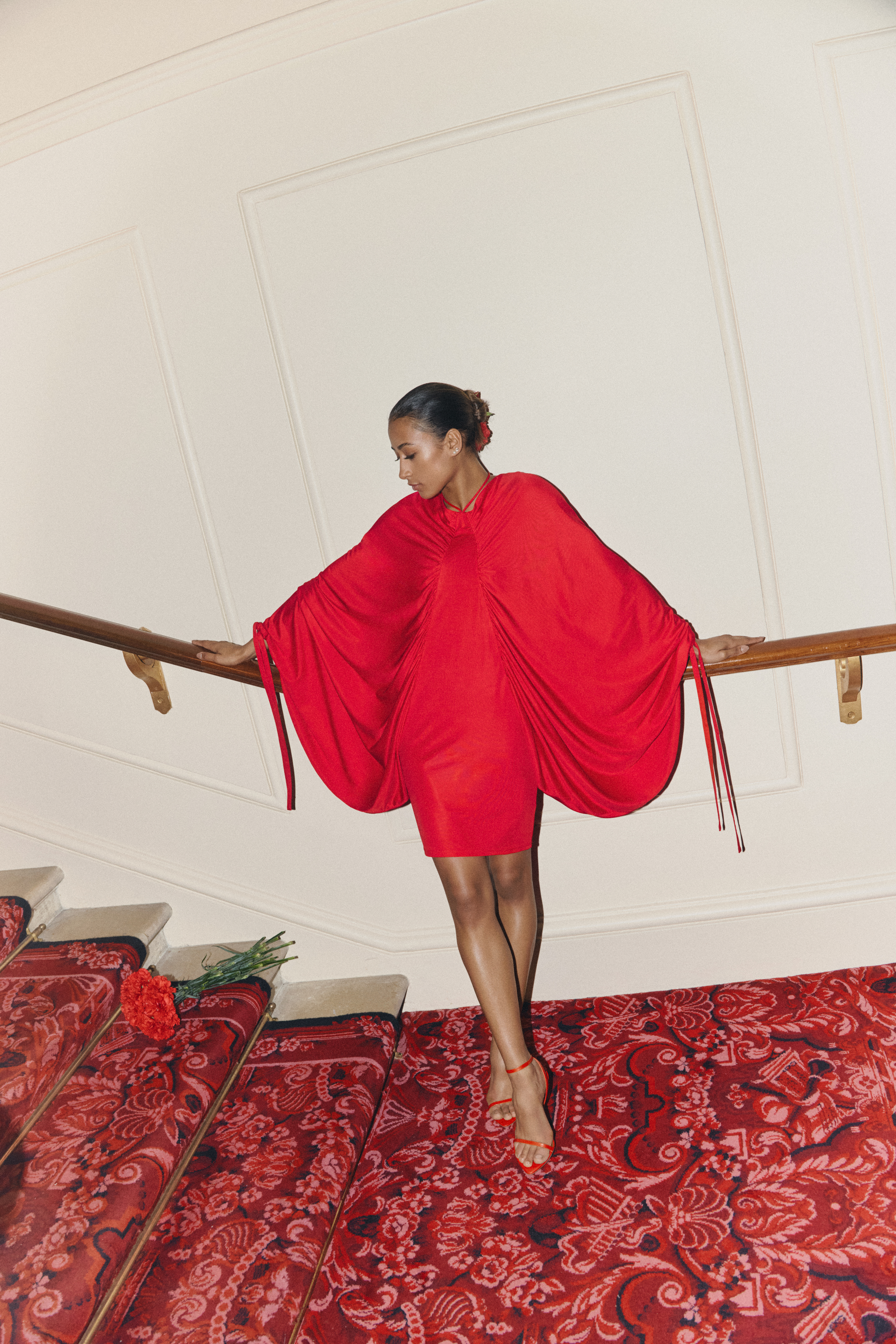 A woman in a red dress with big sleeves rests on a staircase. There are a bunch of red roses on a step to her left.