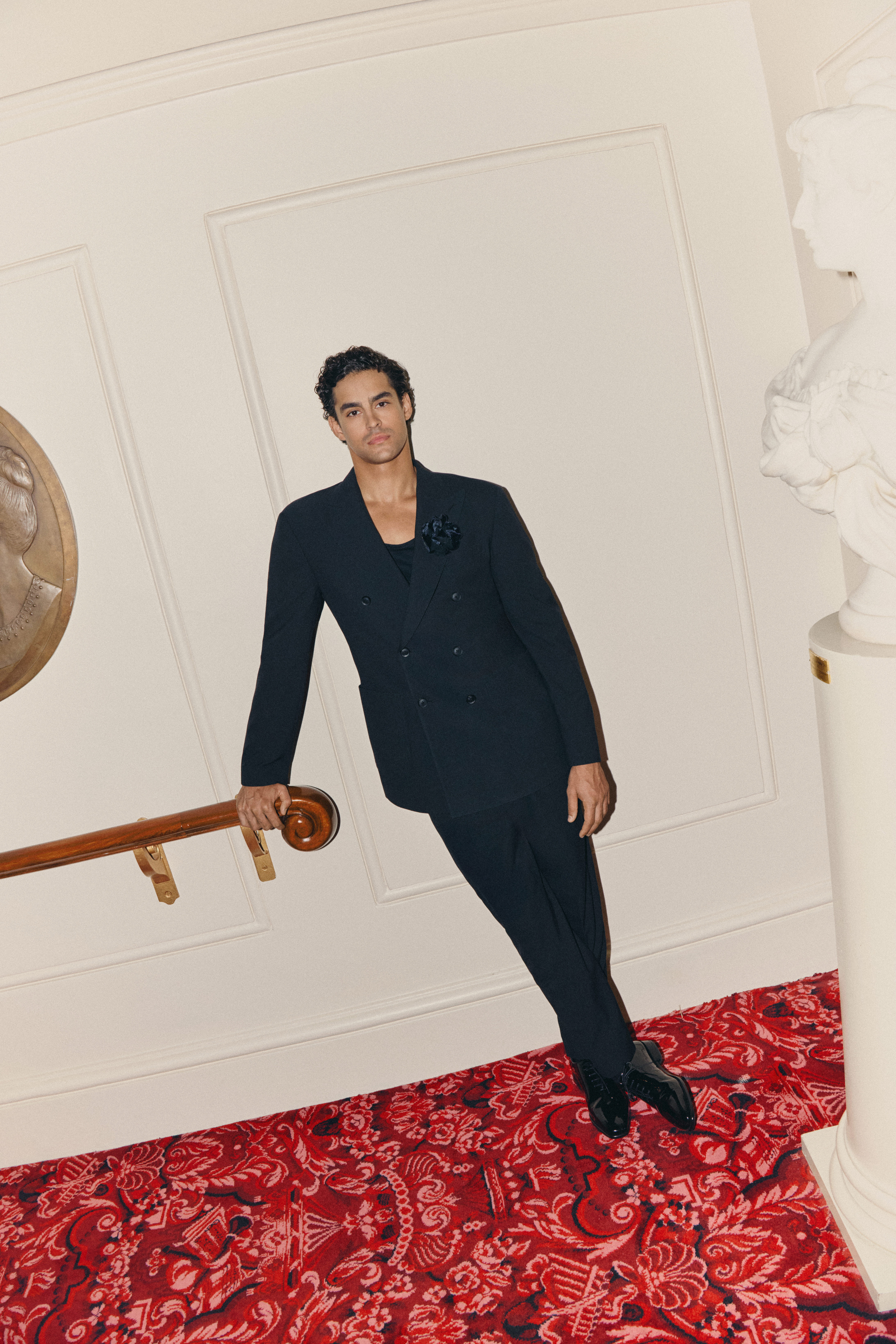 A man in a black double-breasted suit leans on a handrail.