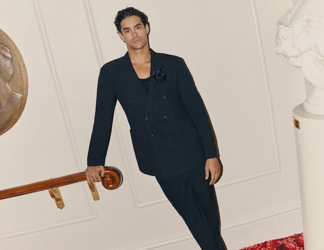 A man in a black double-breasted suit leans on a handrail.