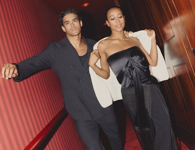A woman in a black satin jumpsuit and white blazer is accompanied by a man in a black suit, walking down a red corridor.