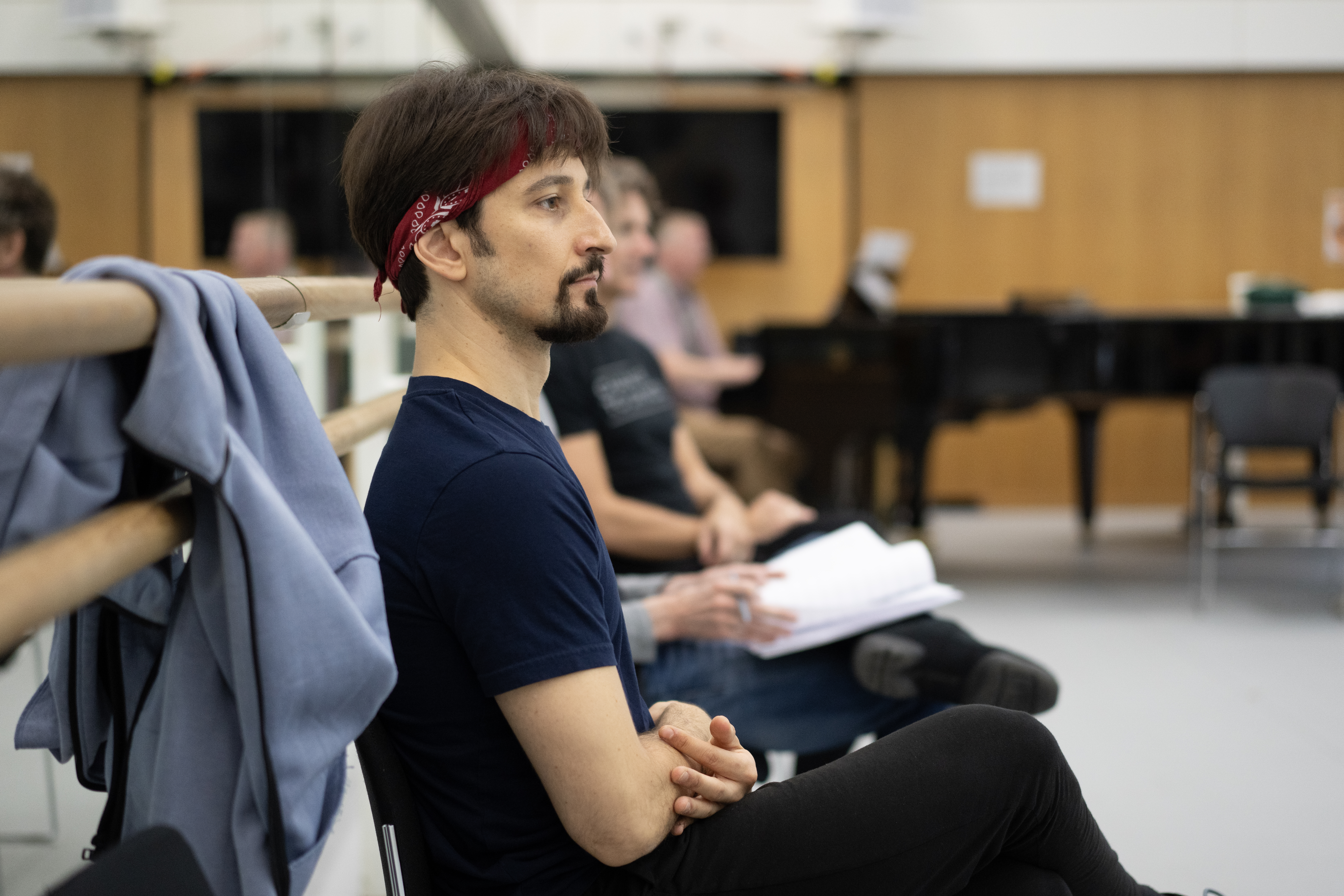 A man with a beard and long dark hair sits on a chair, watching a rehearsal. He is wearing a red bandana in his hair and there is a blue jumper on the barre to his left. (1 of 3)