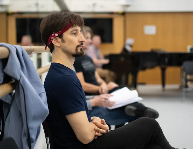 A man with a beard and long dark hair sits on a chair, watching a rehearsal. He is wearing a red bandana in his hair and there is a blue jumper on the barre to his left. (1 of 3)