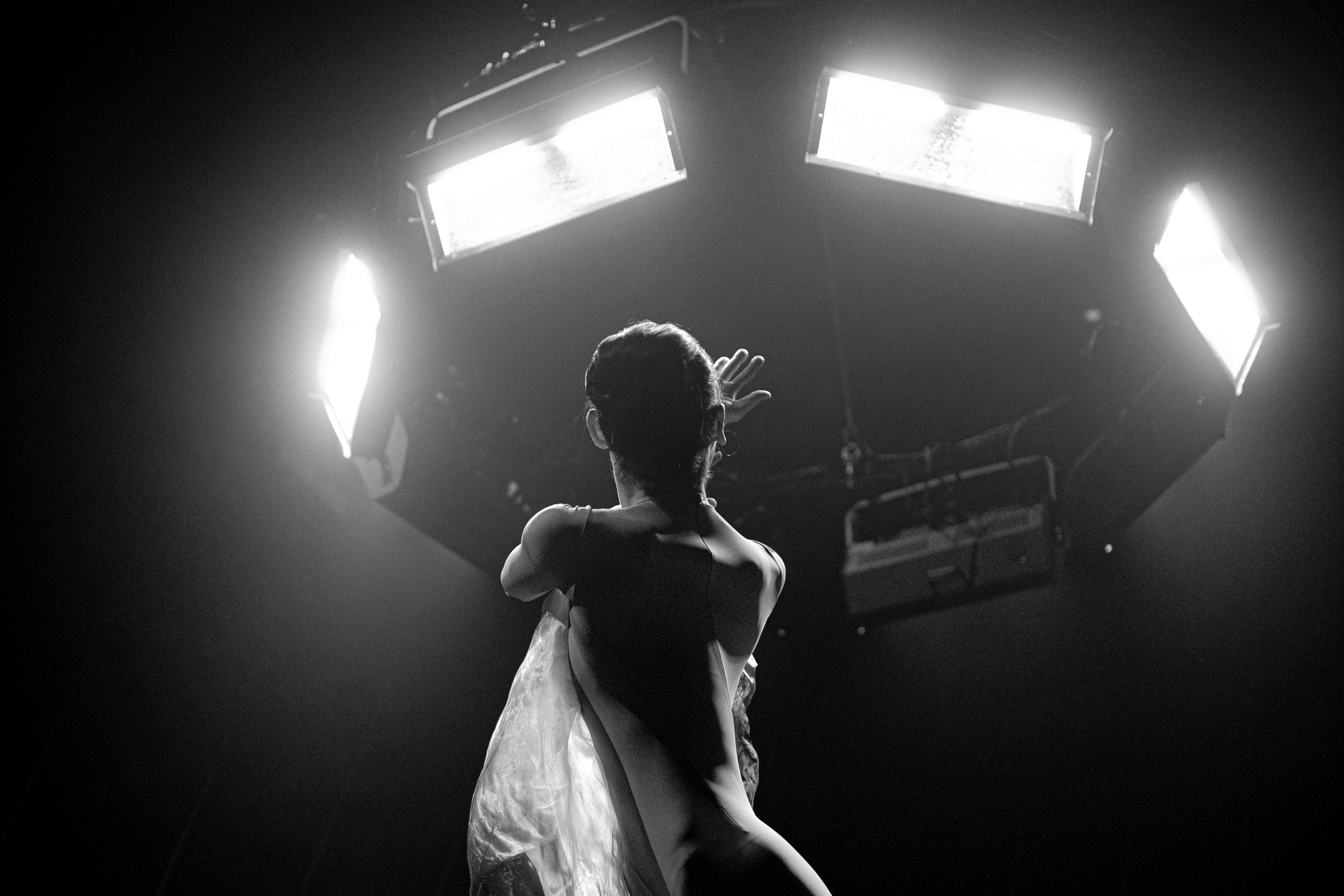 A ballet dancer looks upwards towards rigged lighting with their left arm across their body and their right arm holding on to their torso. The image is in black and white making it hard to decipher details.