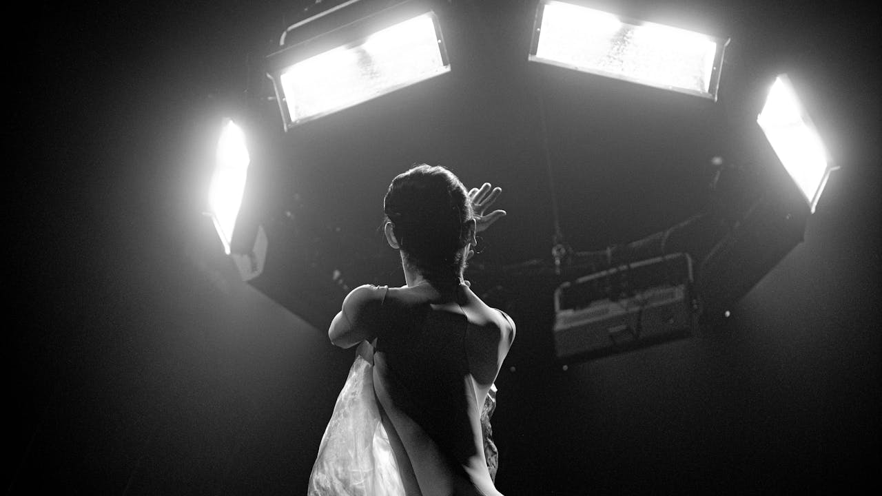 A black and white photo from The Royal Ballet's production of Dark With Excessive Bright. A dancer stands with her back to the camera, she is looking up. Above her a bank of television monitors glow white.
