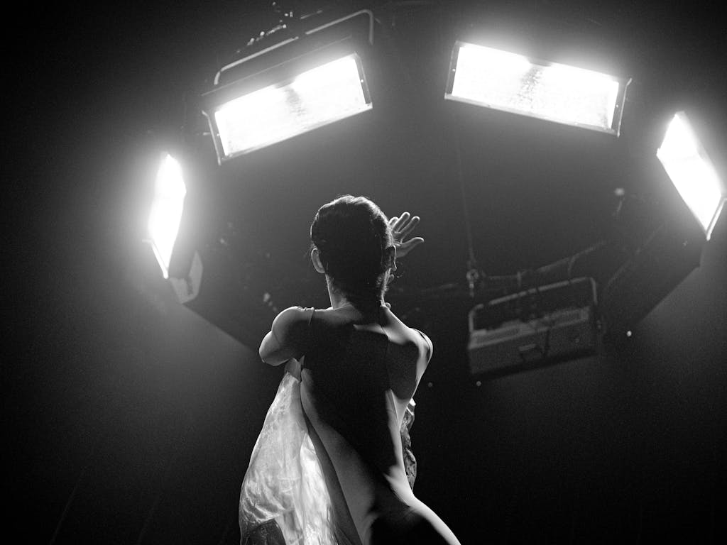 A ballet dancer looks upwards towards rigged lighting with their left arm across their body and their right arm holding on to their torso. The image is in black and white making it hard to decipher details.