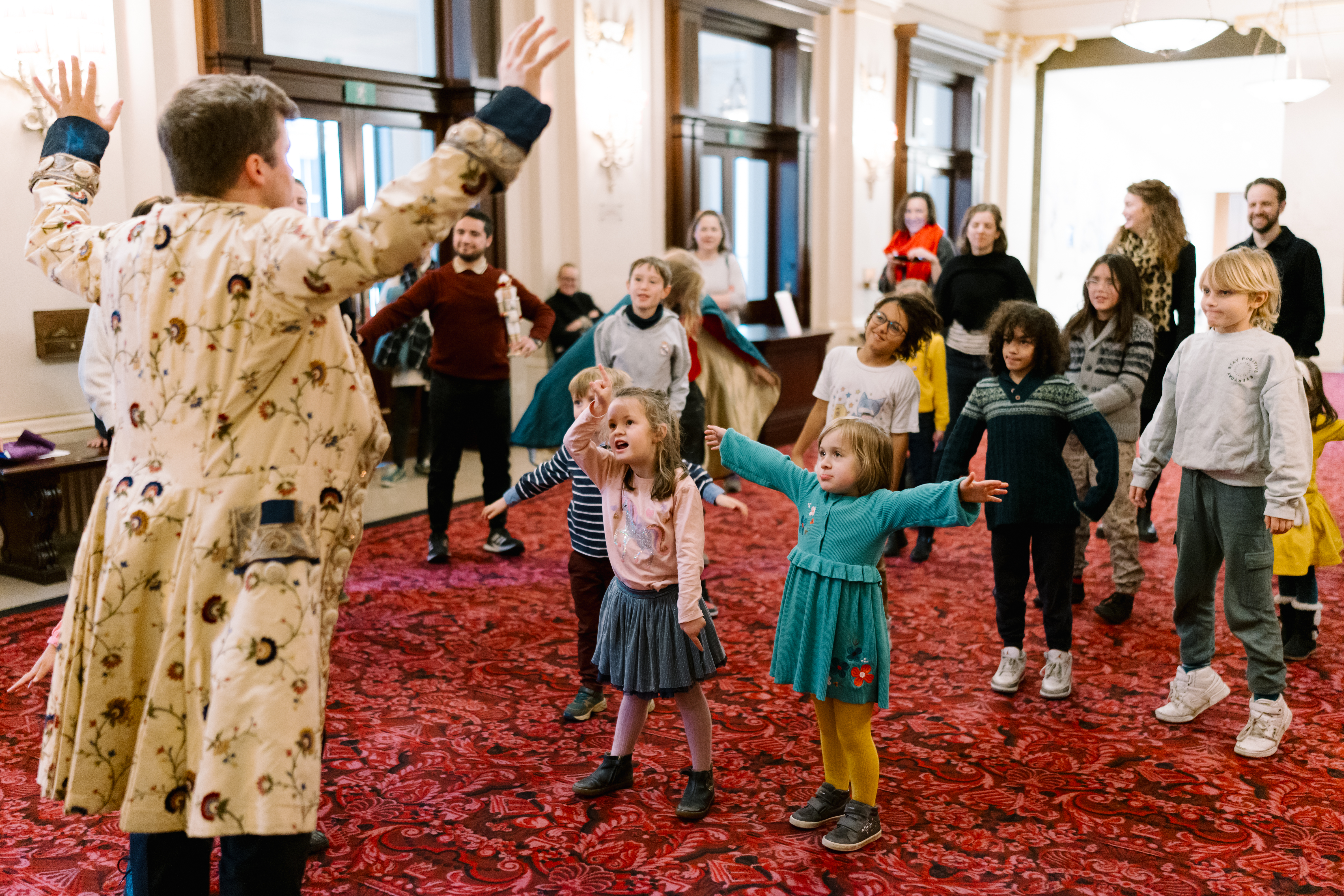 A man in a patterned coat stands with arms outstretched, addressing a group of adults and children in an open indoor space. The group appears engaged, copying the man's action on a red carpeted floor with doors in the background.