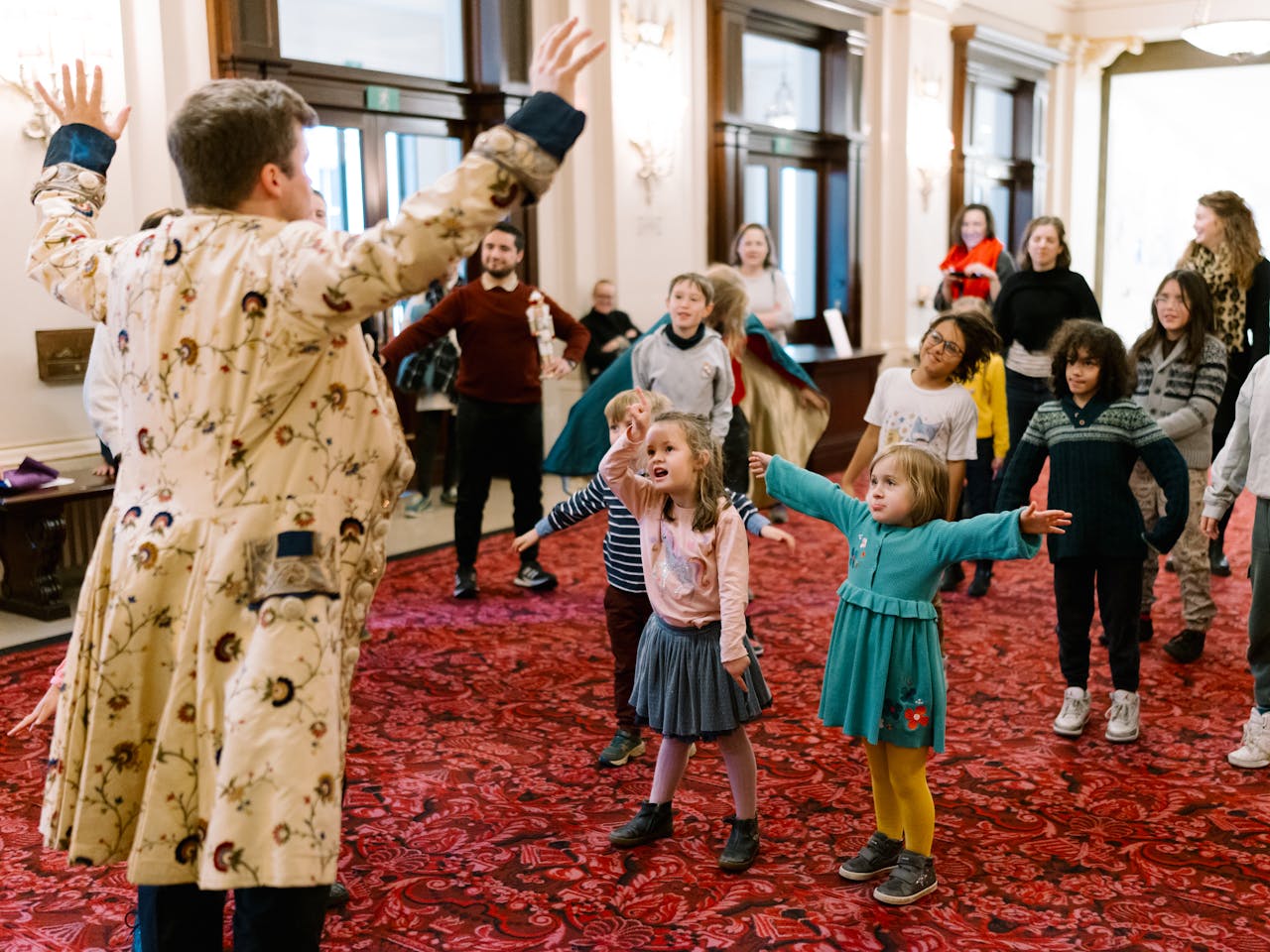 A man in a patterned coat stands with arms outstretched, addressing a group of adults and children in an open indoor space. The group appears engaged, copying the man's action on a red carpeted floor with doors in the background.