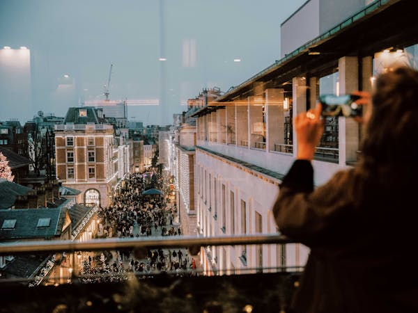 A person takes a photo of a bustling, festive city street from a rooftop. The street is lit with holiday lights, and a large crowd gathers below.