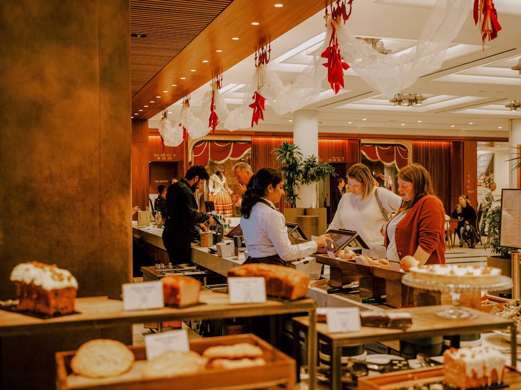A warmly lit cafe with customers browsing and ordering pastries from a server. The ceiling is decorated with red and white drapery, creating a festive atmosphere.