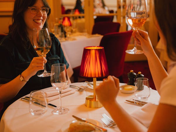 Two people sit at a circular table and raise wine glasses over a white tablecloth. A small red lamp adds warmth.