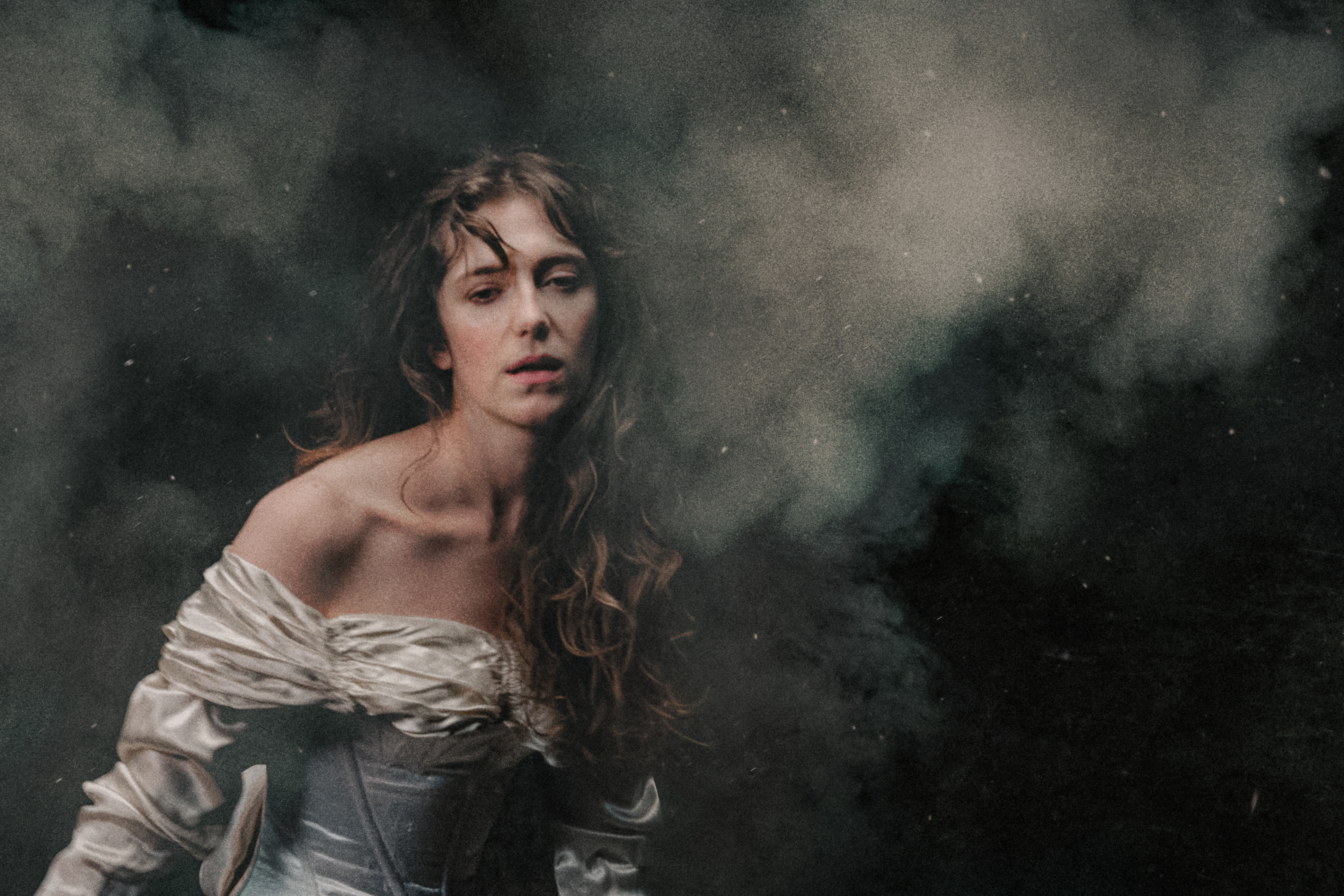 A white woman in an off-the-shoulder, ivory satin gown stands against a dark, smoky background. Her long, wavy hair falls loosely over her shoulders, and she gazes softly forward with a pensive and exhausted expression, illuminated by gentle, diffused light. The air is thick with what looks like battle smoke. The woman represents Elvira in Richard Jones’ production of I Puritani.