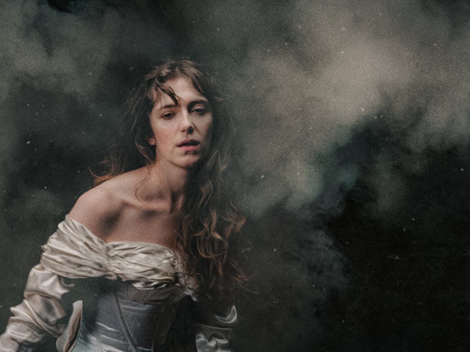 A white woman in an off-the-shoulder, ivory satin gown stands against a dark, smoky background. Her long, wavy hair falls loosely over her shoulders, and she gazes softly forward with a pensive and exhausted expression, illuminated by gentle, diffused light. The air is thick with what looks like battle smoke. The woman represents Elvira in Richard Jones’ production of I Puritani.