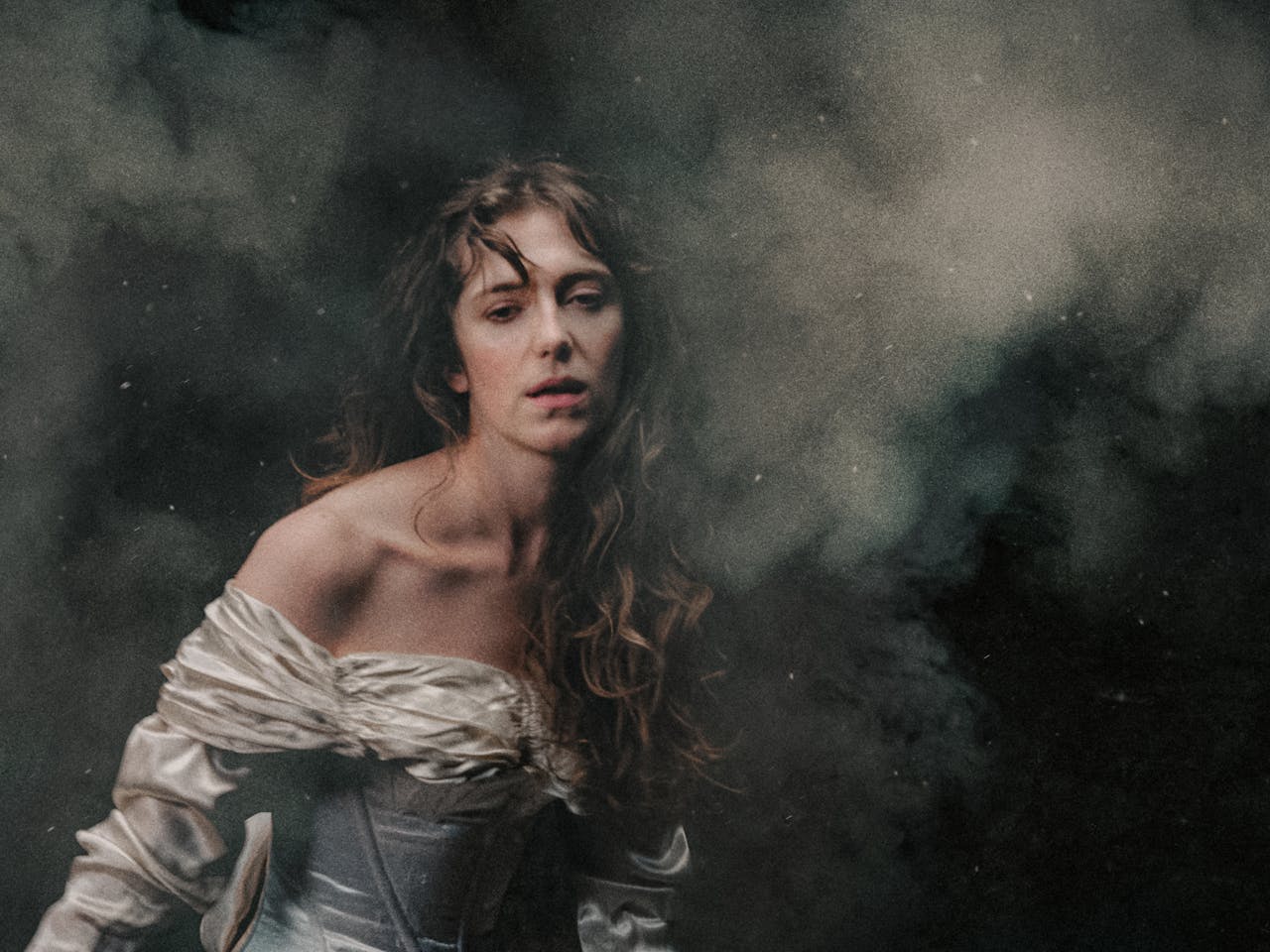 A white woman in an off-the-shoulder, ivory satin gown stands against a dark, smoky background. Her long, wavy hair falls loosely over her shoulders, and she gazes softly forward with a pensive and exhausted expression, illuminated by gentle, diffused light. The air is thick with what looks like battle smoke. The woman represents Elvira in Richard Jones’ production of I Puritani.