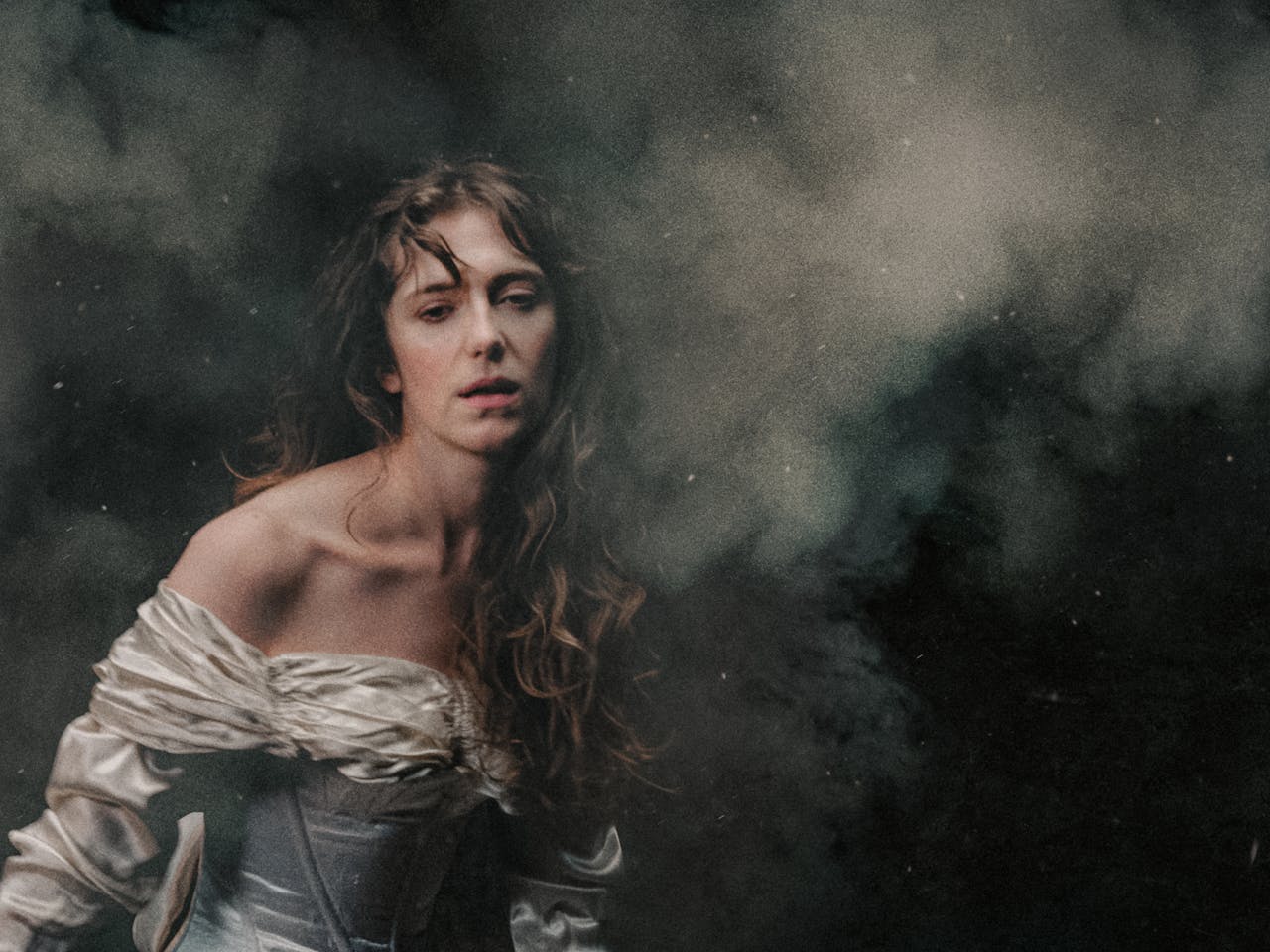 A white woman in an off-the-shoulder, ivory satin gown stands against a dark, smoky background. Her long, wavy hair falls loosely over her shoulders, and she gazes softly forward with a pensive and exhausted expression, illuminated by gentle, diffused light. The air is thick with what looks like battle smoke. The woman represents Elvira in Richard Jones’ production of I Puritani.
