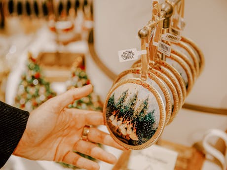 A hand reaches for a festive ornament featuring ballet dancers and snowy trees. The ornaments are hung on a shop display rail.