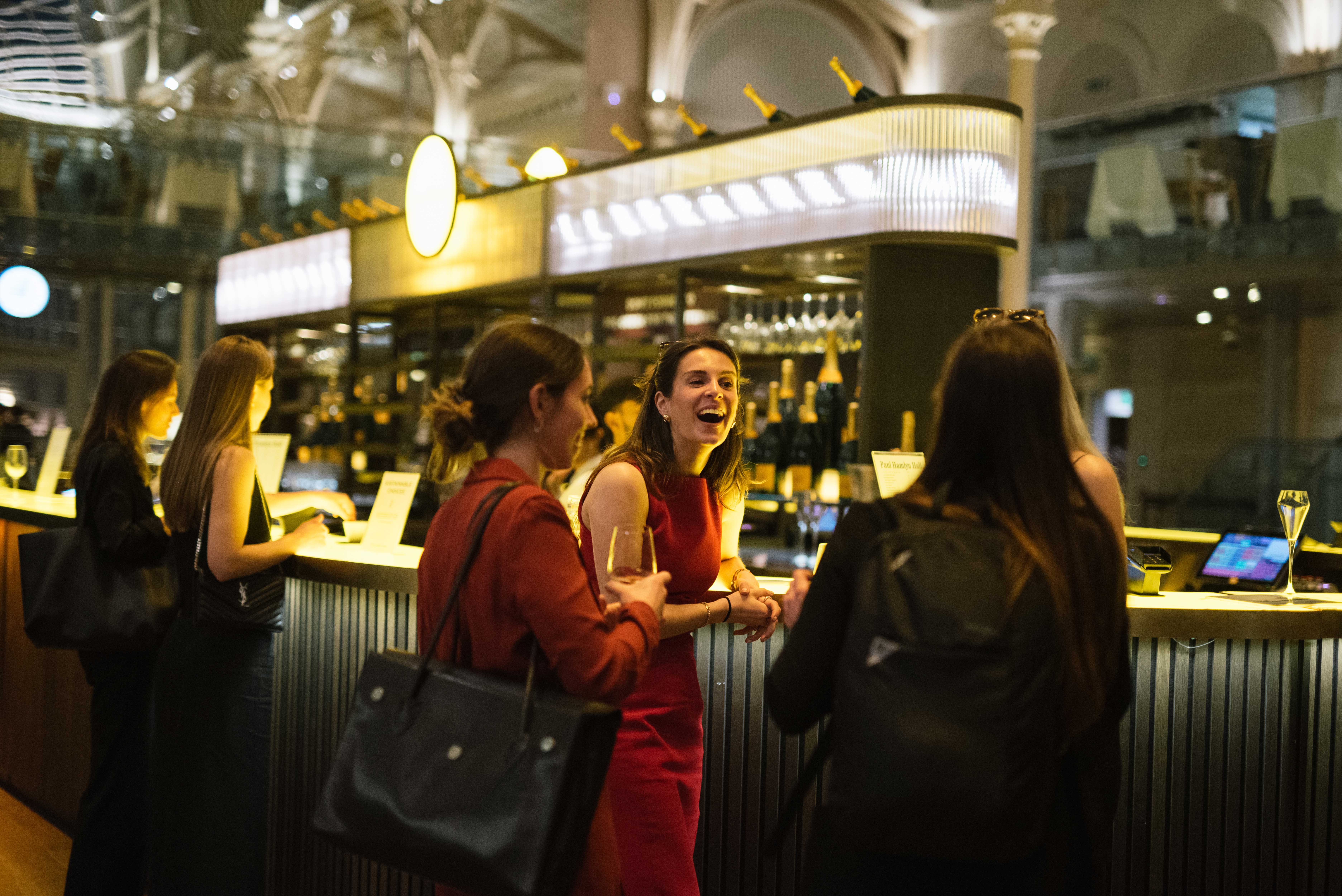 A lively bar scene with five women conversing and laughing, holding drinks. They stand in front of the Veuve Cliquot Champagne Bar in the Paul Hamlyn Hall.