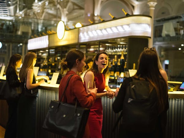 A lively bar scene with five women conversing and laughing, holding drinks. They stand in front of the Veuve Cliquot Champagne Bar in the Paul Hamlyn Hall.
