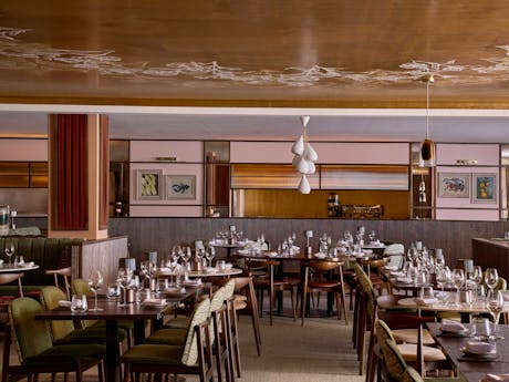 Spacious, elegant restaurant interior with neatly arranged wooden tables and green chairs. Warm lighting and decorative ceiling.