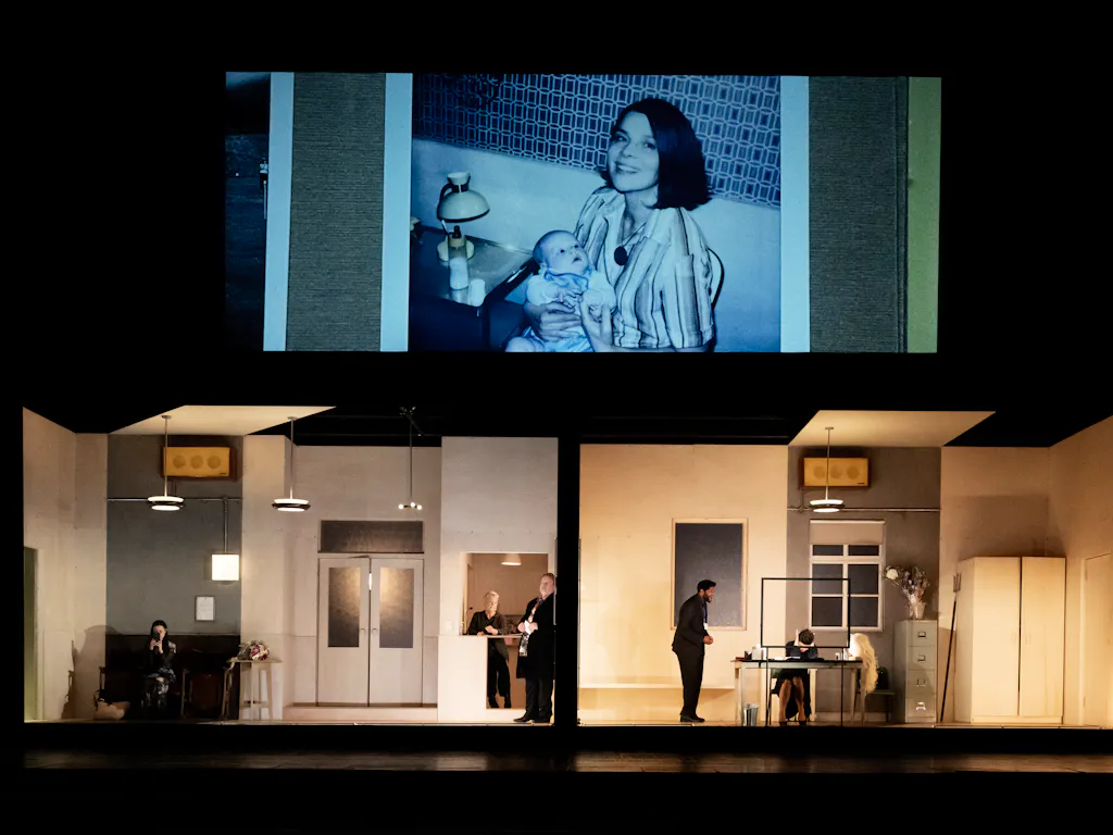 The stage is split into two rooms. Above the rooms is a screen. On the screen is a photo of a dark haired woman smiling and holding a baby.