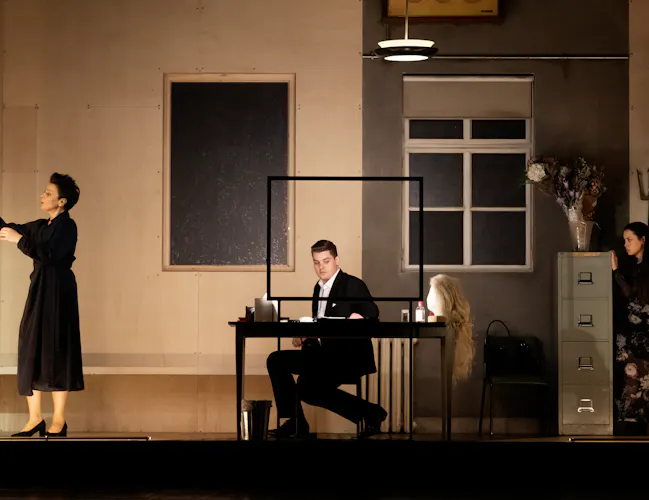 A woman in a black dress hangs her dressing gown up. A man sits at a dressing table. Behind a filing cabinet another woman watches.