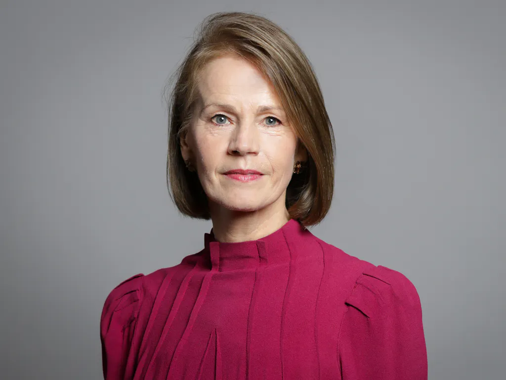 A portrait of Baroness Deborah Bull CBE. She has short brown hair and is wearing a dark pink blouse. She is standing against a grey background.