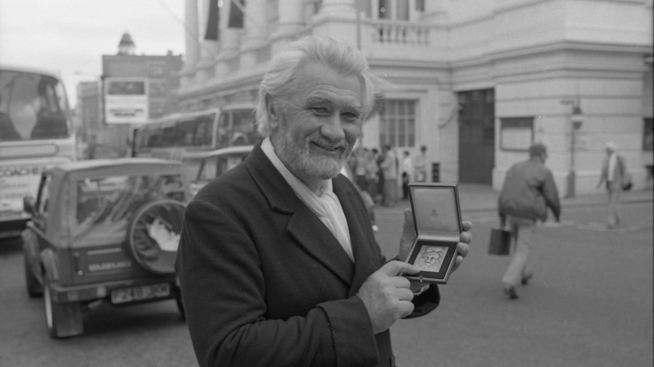 A black and white photo of bass-baritone Donald McIntyre standing in front of the Royal Opera House. He wears a suit and holds a medal in his hand.