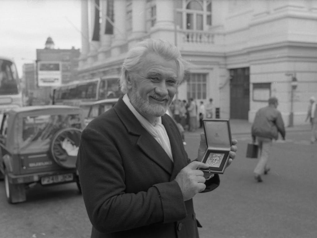 A black and white photo of bass-baritone Donald McIntyre standing in front of the Royal Opera House. He wears a suit and holds a medal in his hand.