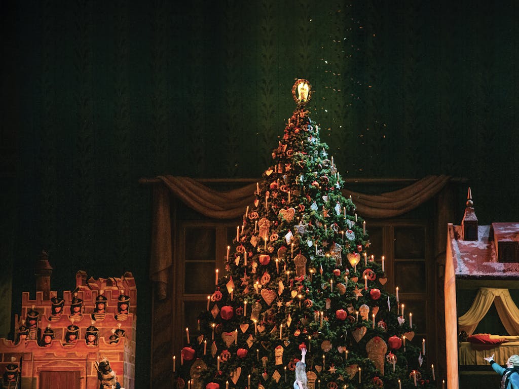 A photo of the Royal Opera House Main Stage during a production of The Nutcracker. A large, beautifully decorated Christmas tree stands in the middle of the stage. Clara, dressed in white, stands in front of the tree with her back to the audience and points up in amazement. To the side Herr Drosselmeyer looks on in a blue cape and also points at the Christmas tree.
