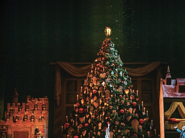 A photo of the Royal Opera House Main Stage during a production of The Nutcracker. A large, beautifully decorated Christmas tree stands in the middle of the stage. Clara, dressed in white, stands in front of the tree with her back to the audience and points up in amazement. To the side Herr Drosselmeyer looks on in a blue cape and also points at the Christmas tree.