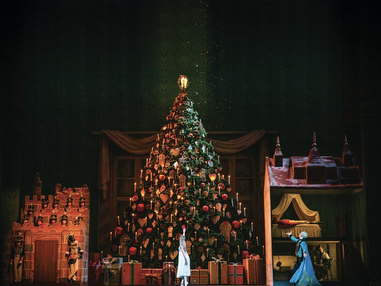 A photo of the Royal Opera House Main Stage during a production of The Nutcracker. A large, beautifully decorated Christmas tree stands in the middle of the stage. Clara, dressed in white, stands in front of the tree with her back to the audience and points up in amazement. To the side Herr Drosselmeyer looks on in a blue cape and also points at the Christmas tree.